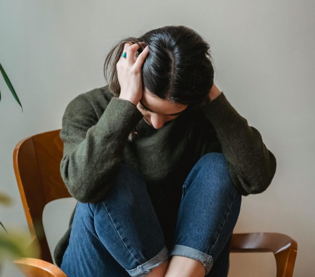 A young woman sitting on a chair indoors, holding her head in distress.